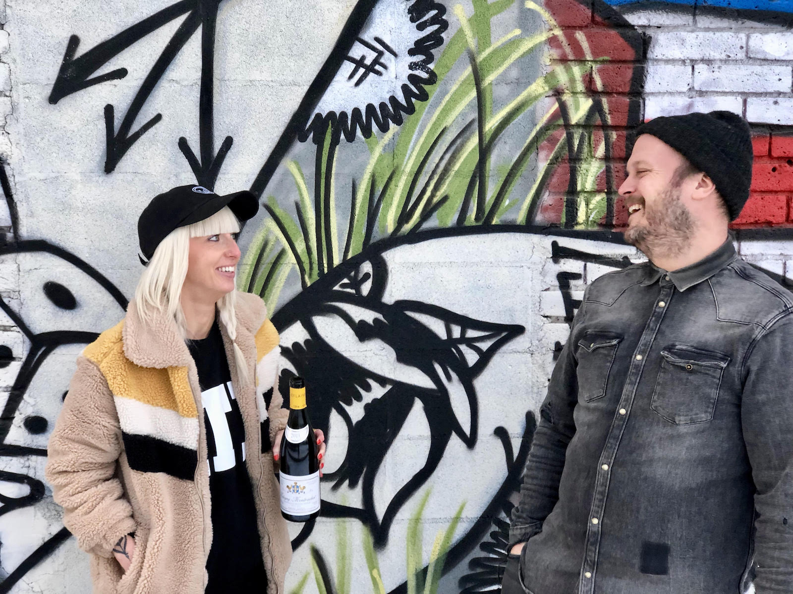 A woman in a winter coat and hat faces a man in a dark gray shirt in front of a graffiti mural.