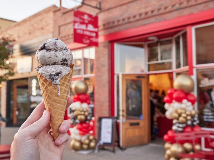 A hand holding a double-scoop ice cream cone in front fo Little Man Ice Cream shop in Littleton