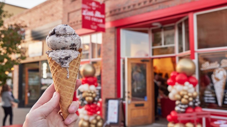 A hand holding a double-scoop ice cream cone in front fo Little Man Ice Cream shop in Littleton