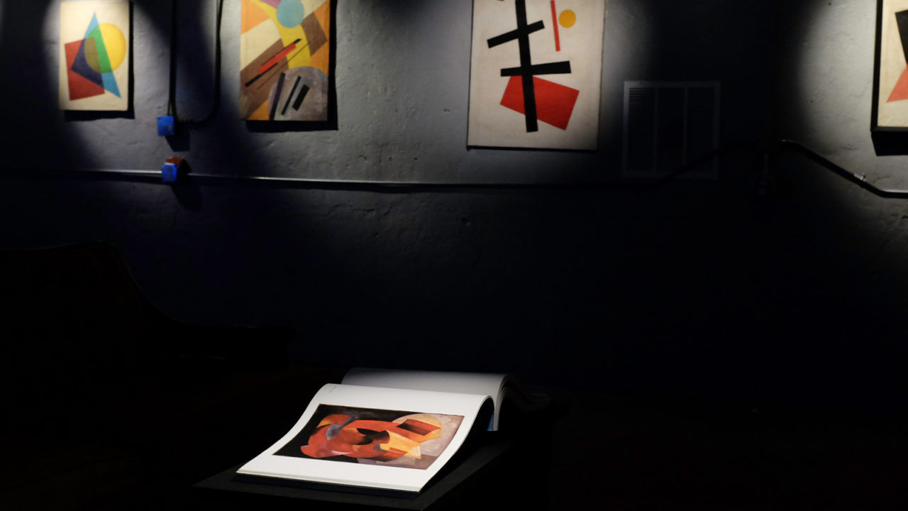 A painting sits in a spotlight in front of four others in a dark room.