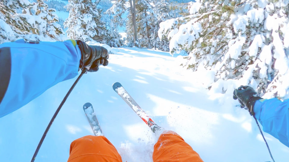 A skier wearing orange pants and a periwinkle parka slashes through powder on a blue-sky day.