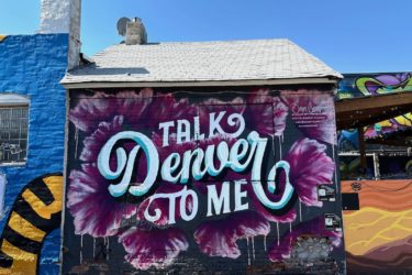 A photo of mural "Talk Denver To Me" by Ashley Joon and Adam Vicarel, featuring the words "Talk Denver To Me" surrounding by purple flowers