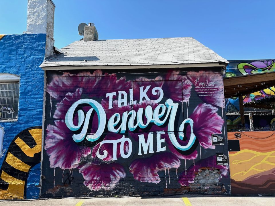 A photo of mural "Talk Denver To Me" by Ashley Joon and Adam Vicarel, featuring the words "Talk Denver To Me" surrounding by purple flowers