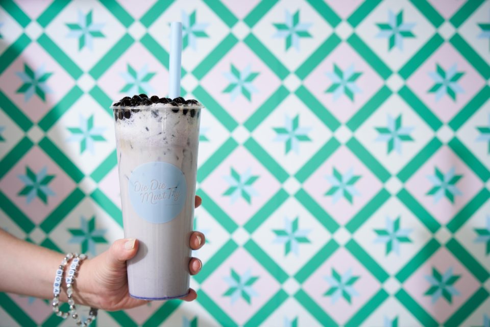 A creamy drink in a tall glass with a straw and a topping of black boba pearls with vibrant blue, pink, and green wallpaper in the background.