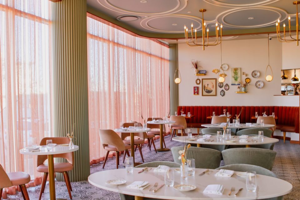 A restaurant dining room with round tables, padded chairs, and a large floor-to-ceiling window with pale pink curtains