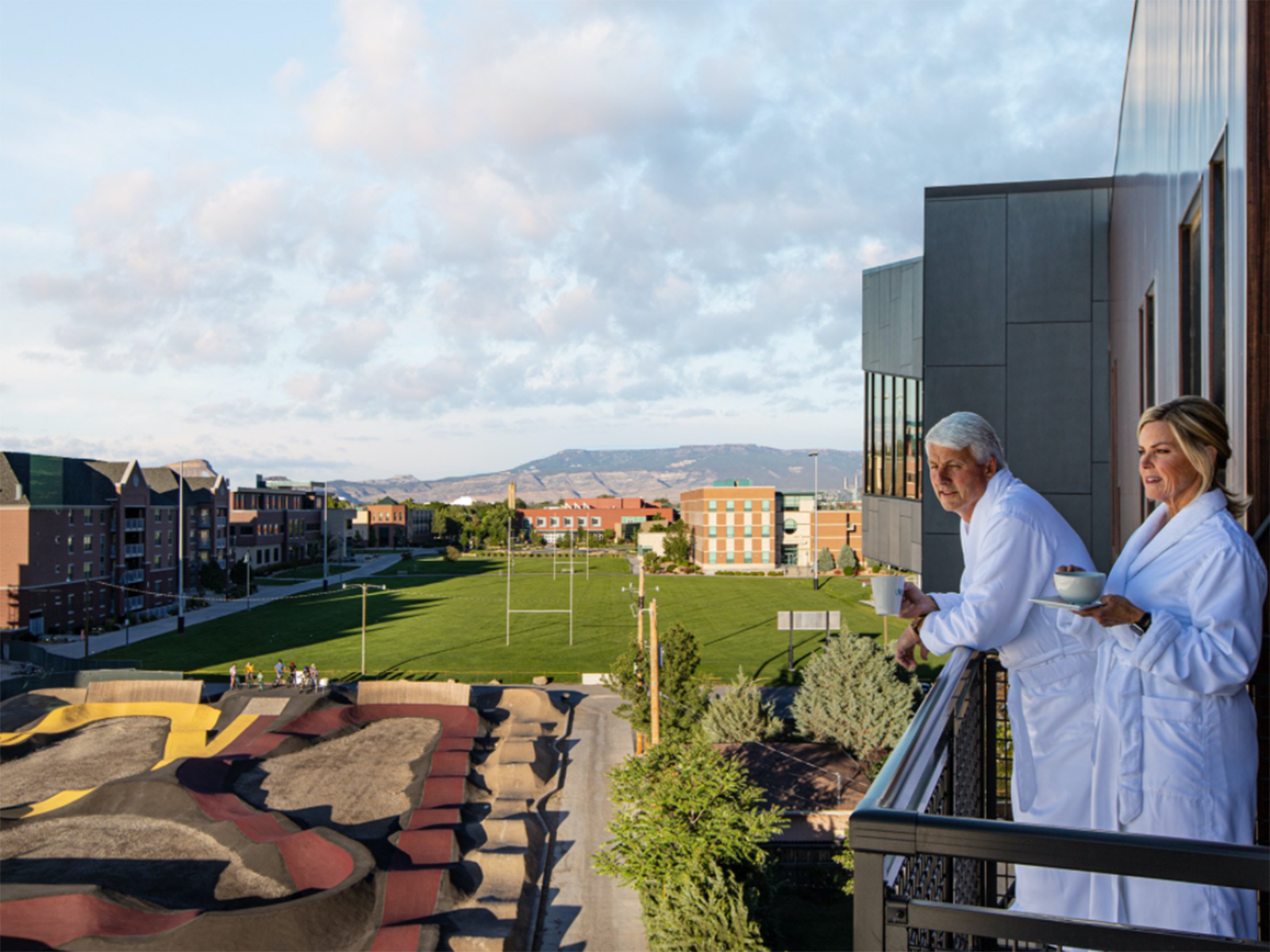 A couple stands on a balcony in white robes watching bikers on a pump track below