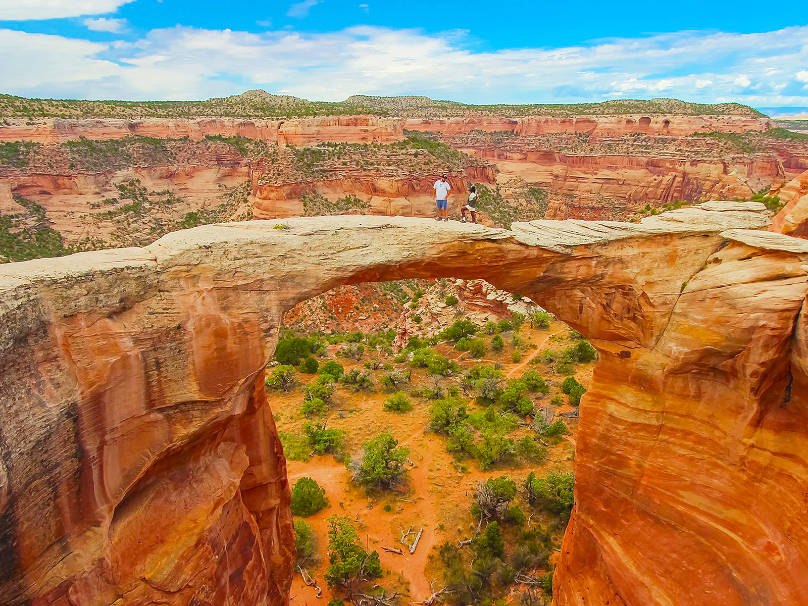 Two people stand atop a natural sandstone arch in a desert