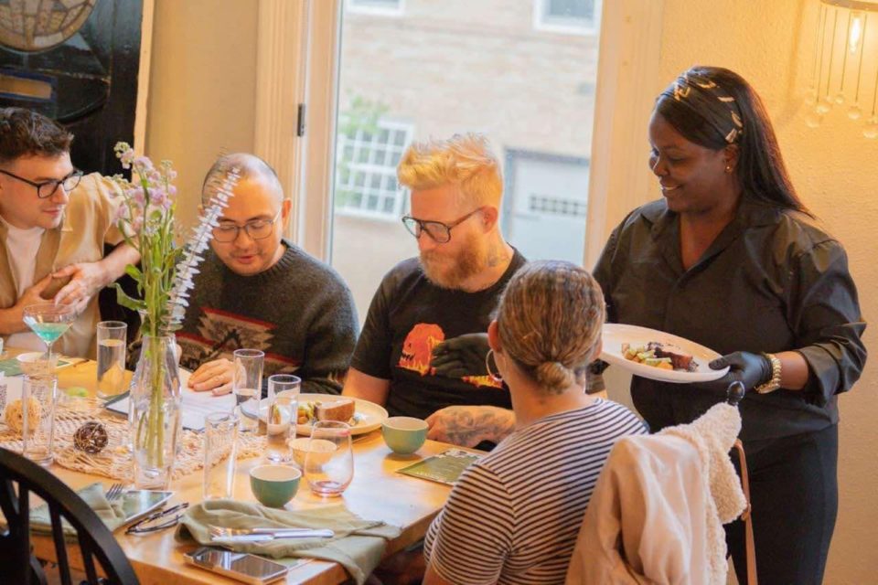 Four people sitting at a table filled with food with a fifth person standing up and presenting another plate of food.