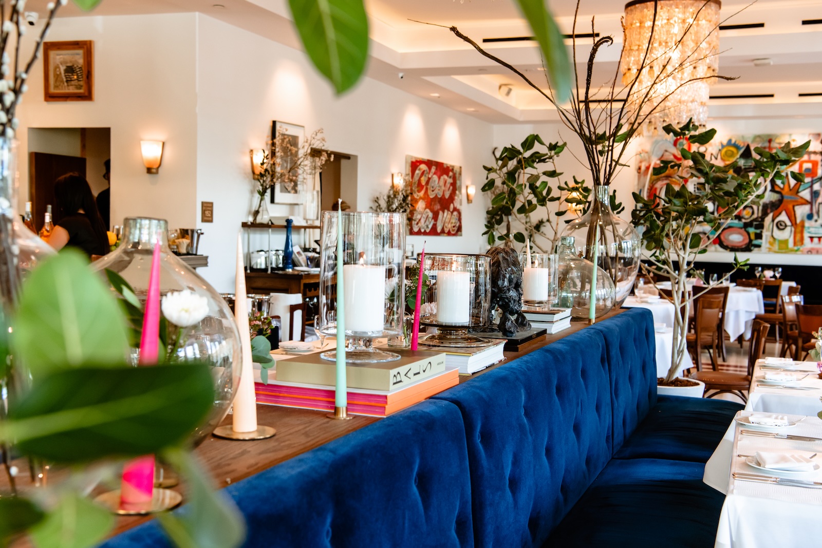A restaurant dining room with a blue velvet banquet, white tableclothes, a running board with books, candles, clear glass vases, and indoor plants