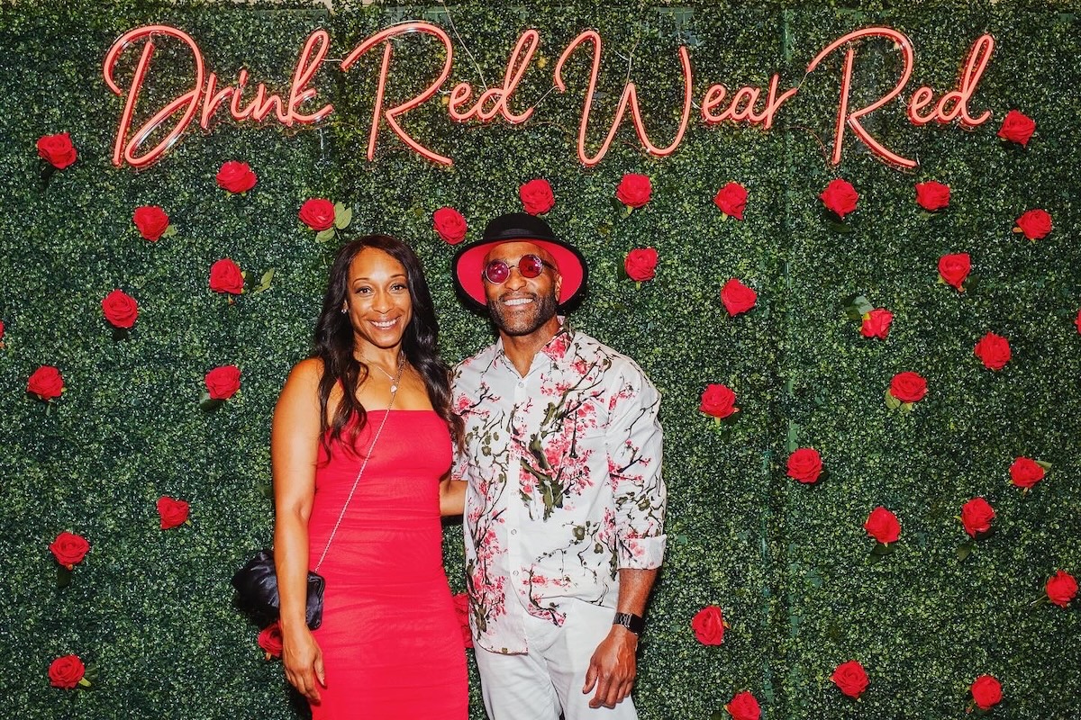 A couple dressed in red stands in front of a wall of flowers and vines with "drink red wear red" in neon