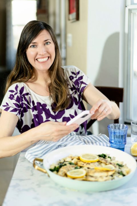 A woman in a purple and white shirt uses her phone to take a picture of a bowl of food.