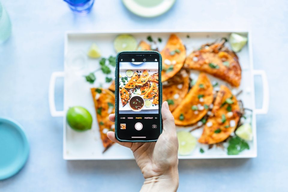 A smart phone taking a picture of a white tray of birria tacos o a white table top