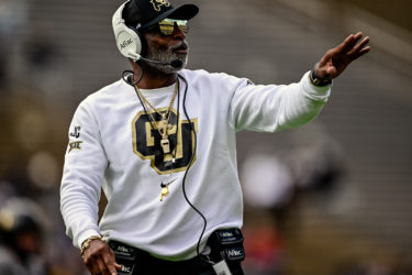 Deion Sanders wears a white CU sweatshirt and coaches on the sideline
