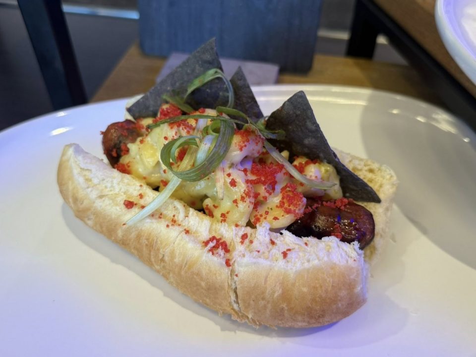 A grilled sausage in a bun with cheese sauce, blue corn tortilla chips, orange sprinkles, and sauce