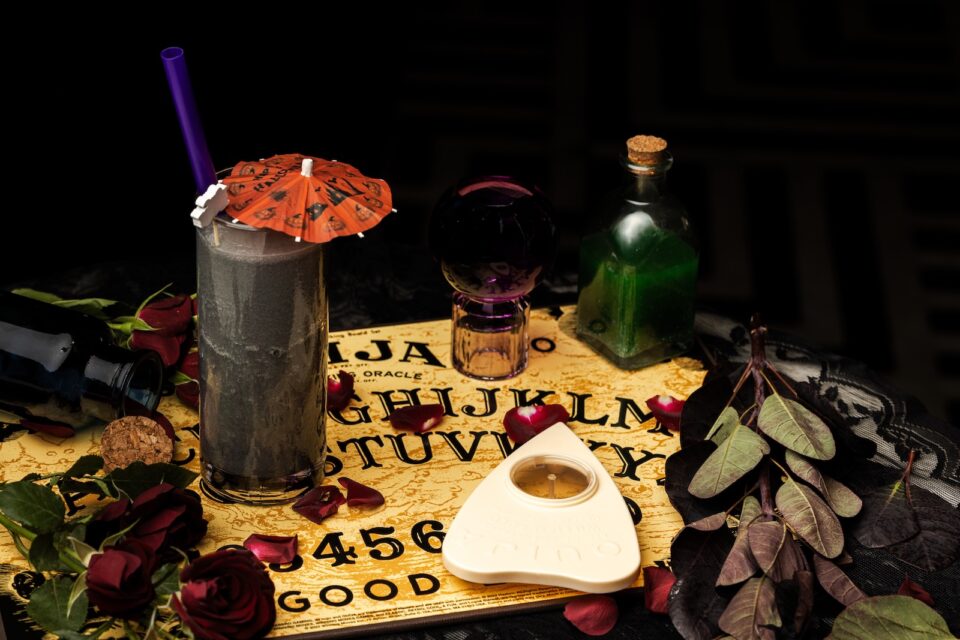 A Ouija board with a black cocktail in a clear glass with a red umbrella.