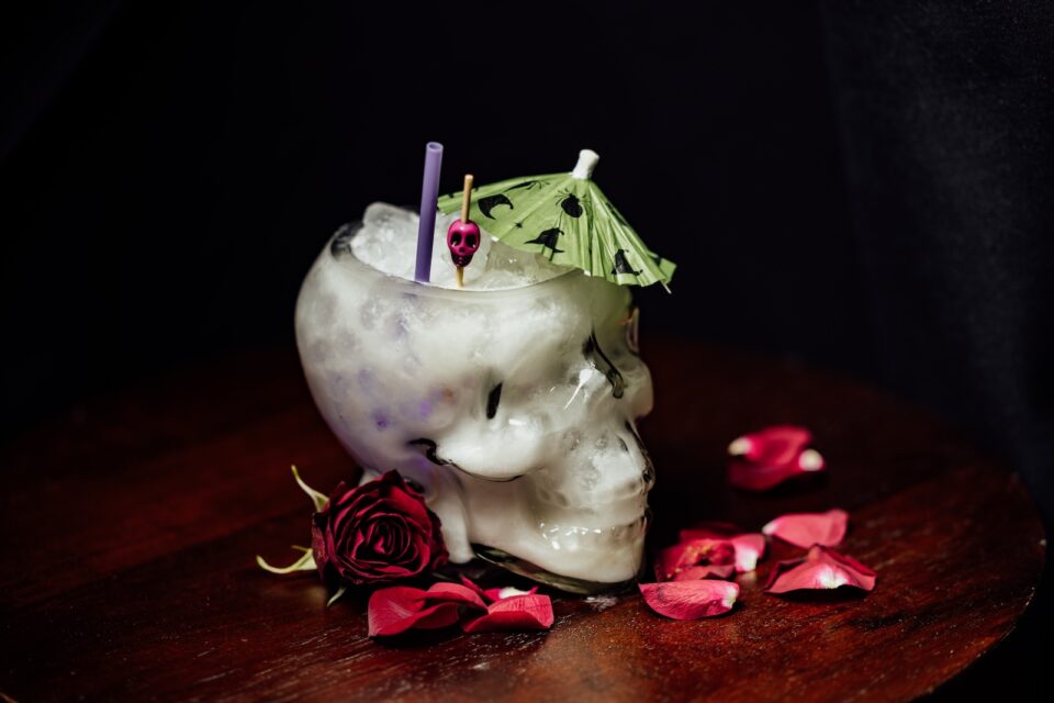 A milky cocktail in a skull-shaped glass with a green umbrell surrounded by rose petals