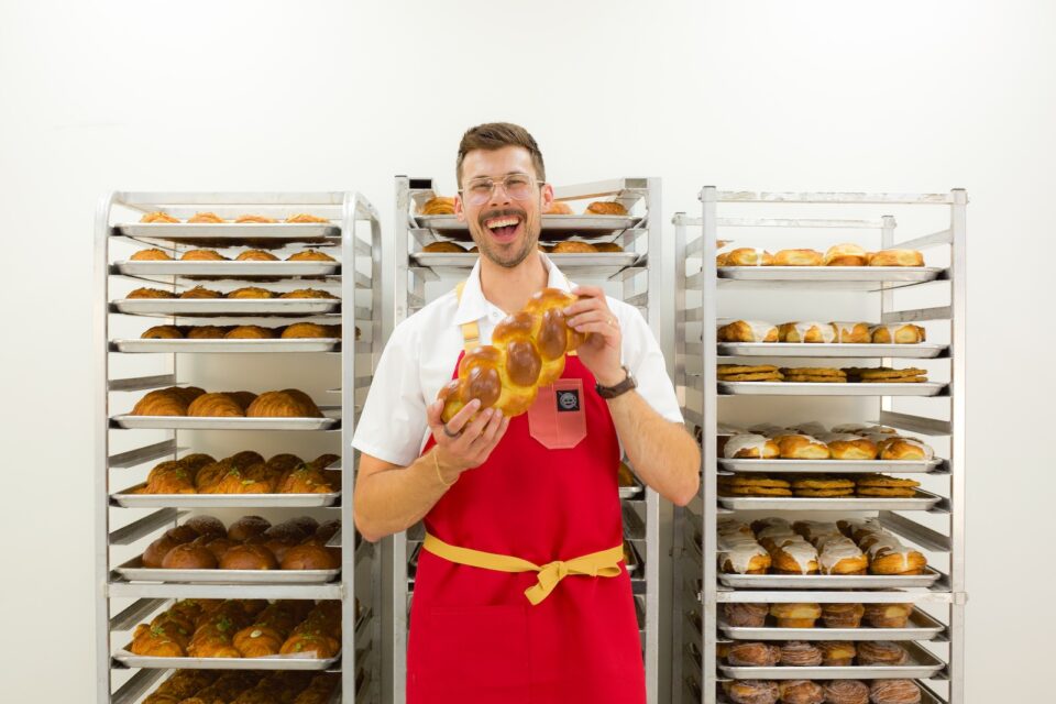 A man with a mustache wearing a red apron and holding a challah loaf in front of racks of baked goods.