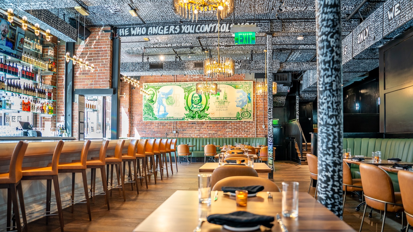 A restaurant interior with tables, leather chairs, a row of bars stools, and a large dollar bill image on a brick wall