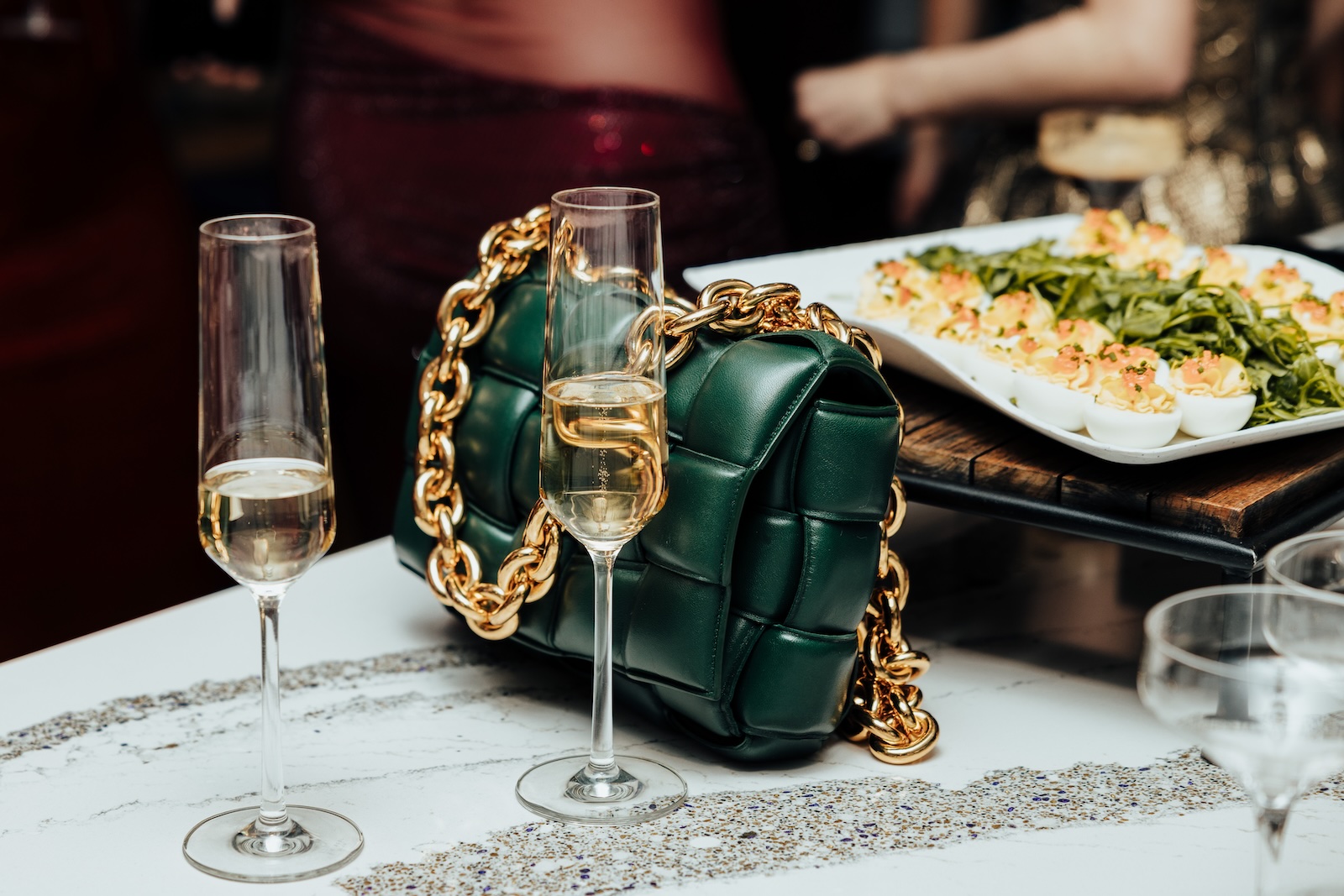 A green purse with a gold chain handle on a table with two champagne glasses and a tray of deviled eggs