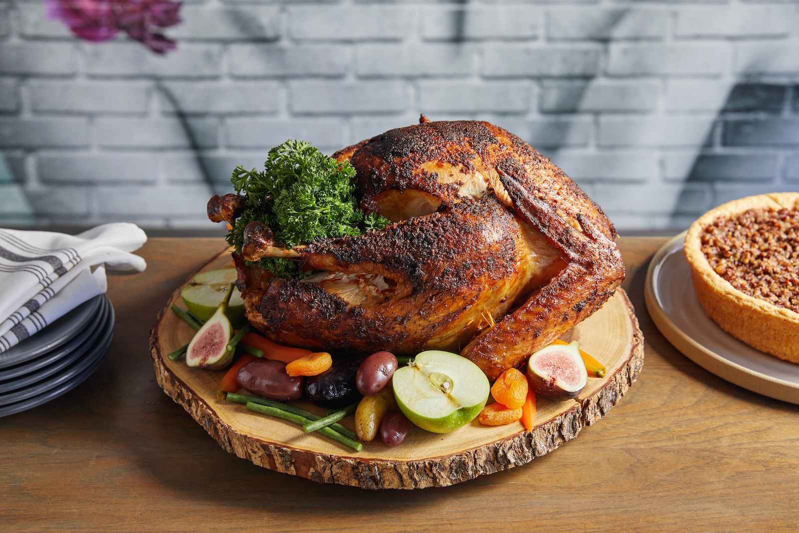 A whole roast turkey on a wooden serving board surrounded by sliced fruits and herbs