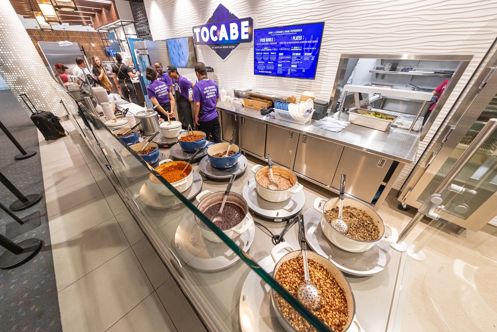 Assorted toppings or sauces in bowls on a counter at a food station, with staff preparing food in the background.
