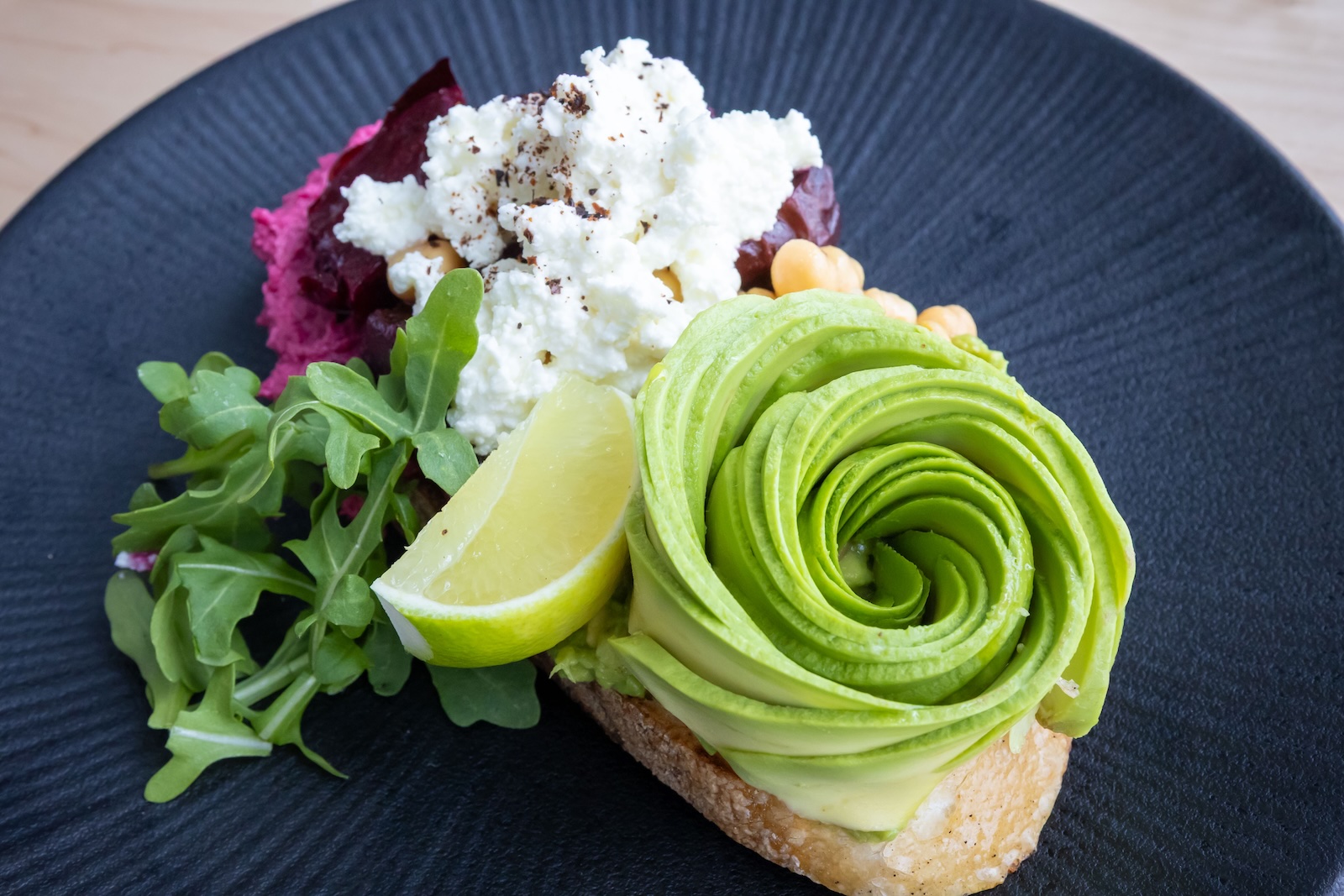 Avocado toast with spiral avocado slices on whole grain bread, served with arugula, lemon wedge, beetroot, and cottage cheese on a black plate.