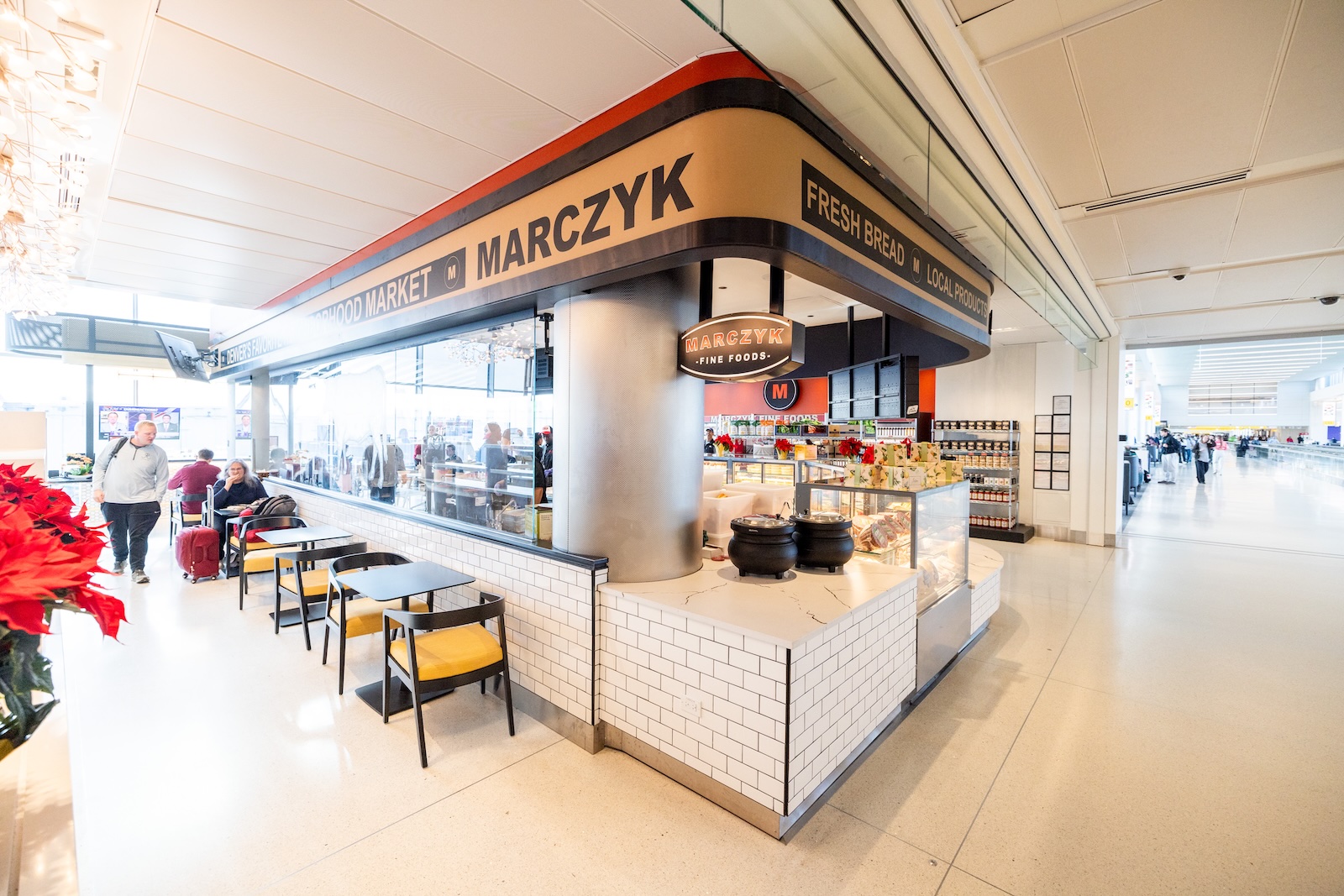 A café at an airport terminal with seating, serving fresh bread, and a sign reading "Marczyk." Passengers walk by in the background.