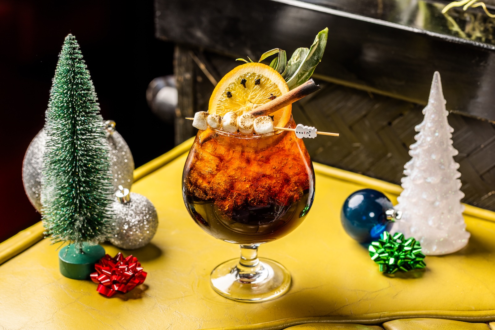 A brown cocktail in a glass goblet garnished with fruit slices and mini toasted marshmallows and surrounded by Christmas ornaments