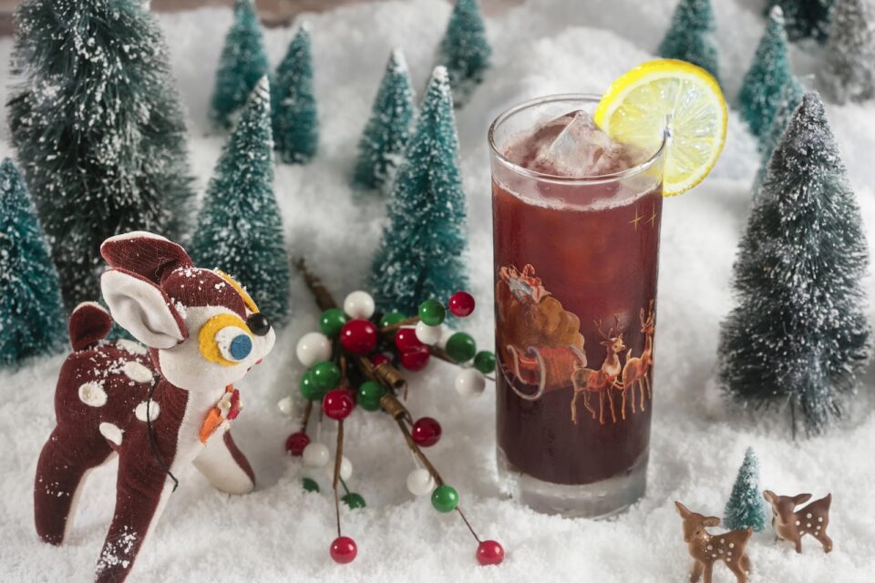 A brown cocktail in a tall glass next to a miniature winter scene with Rudolph the reindeer, pine trees, and snow.