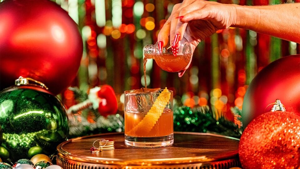 A hand with red nails pouring a drink from a small round flask into a cocktail glass with Christmas decorations all around