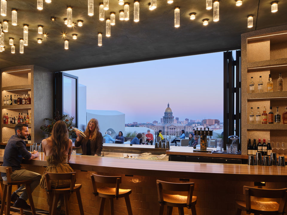 Bartender shakes a drink for a couple sitting at a bar. Through the large window, the Colorado State Capitol is visible.