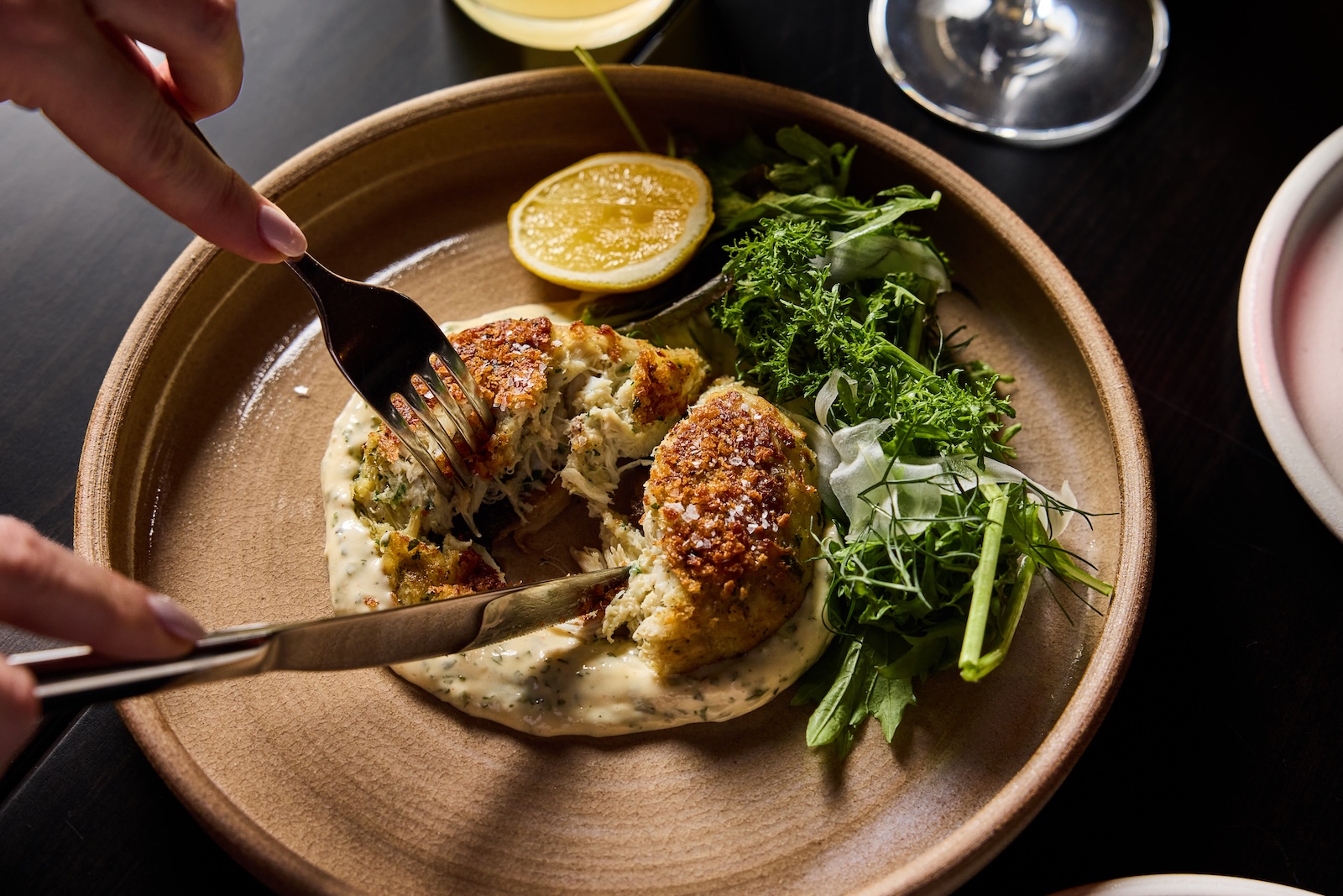 Two hands use a knife and fork to cut into a crab-stuffed mushroom nestled in creamy sauce next to lettuce greens and a lemon wedge on an earthenware plate.