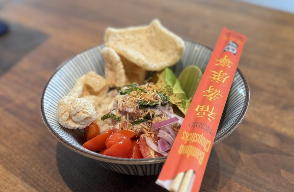 A bowl holding rice topped with marinated fish, vegetables, and pork rinds, with chopsticks in red paper wrapper