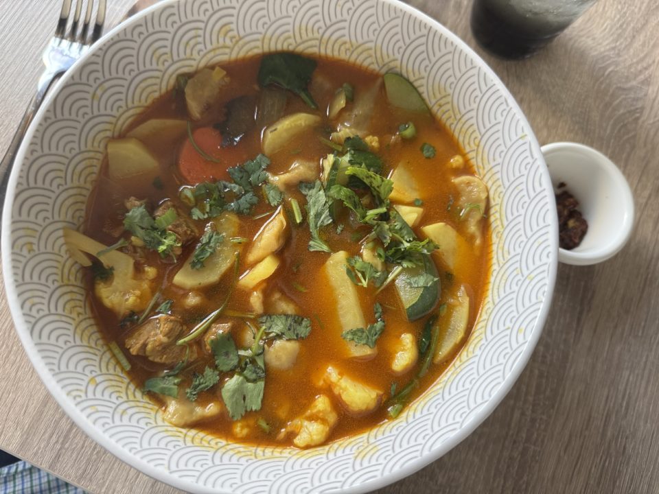 A white bowl holding a tomato broth with vegetables, chunks of lamb, and wheat dumplings.