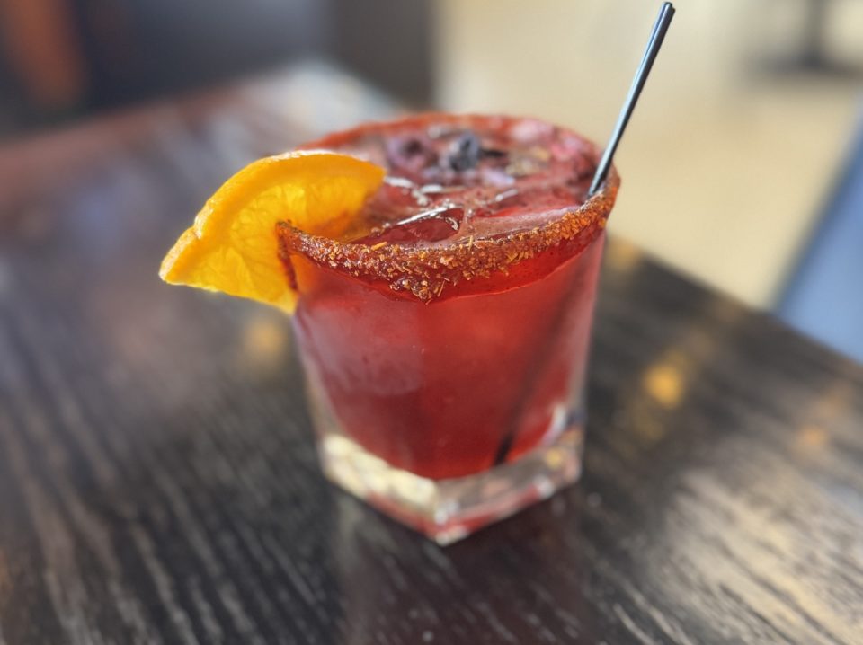 A red cocktail on the rocks with an orange rim and spices on the rim