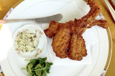 Three deep-fried frog legs on a plate with fresh greens, a small bowl of compound butter, and a butter knife