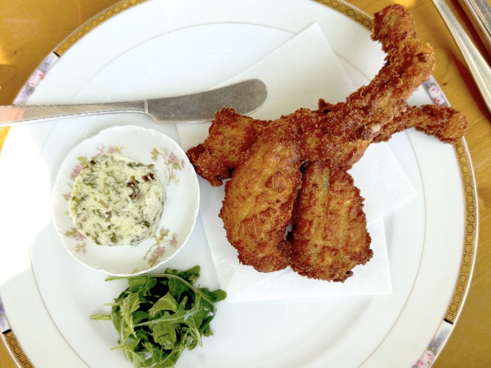Three deep-fried frog legs on a plate with fresh greens, a small bowl of compound butter, and a butter knife