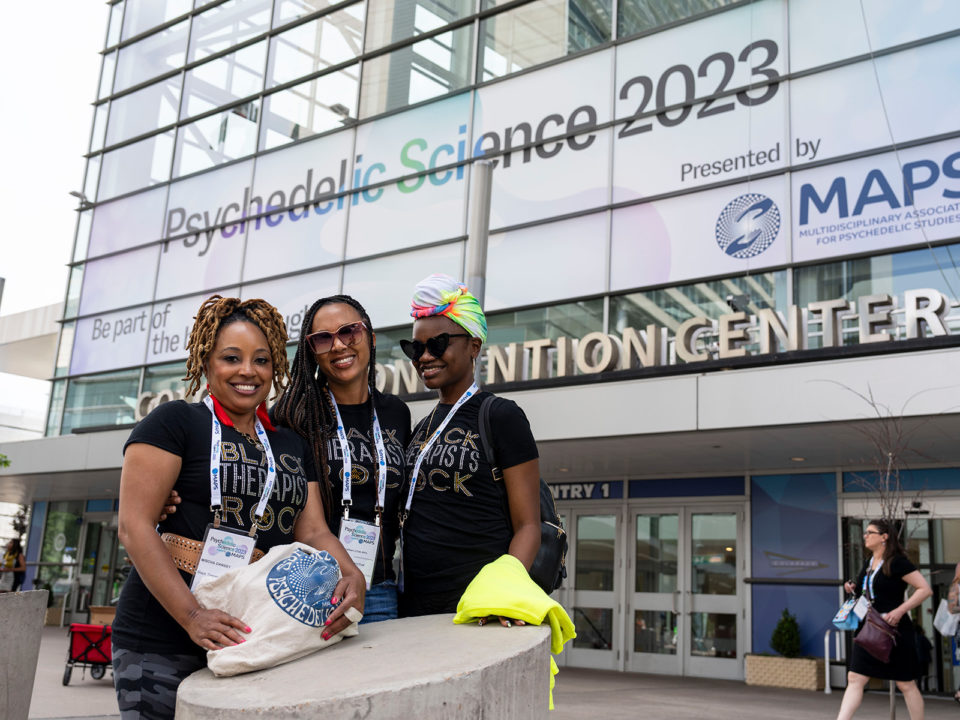 Three black women smile in front of the Psychedelic Science 2023 conference banner