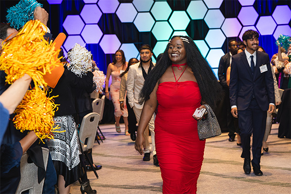 A Black teenage girl wearing a red dress walking down an aisle with other teens walking behind her.