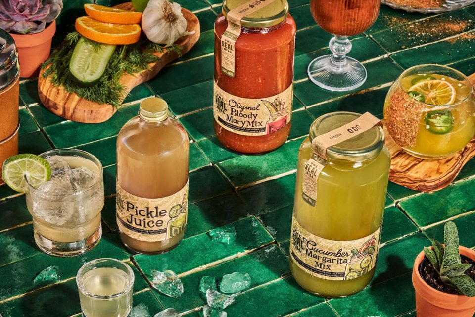 Bottles of the Real Dill cocktail mixes and cocktails on ice in glasses.