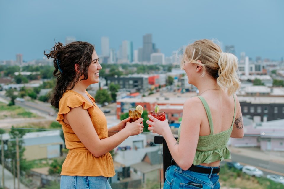 Two women hold cocktails on the edge of a rooftop patio with a downtown skyline in the background.