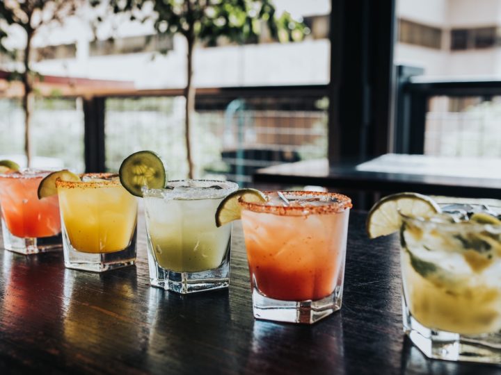 Five glasses of colorful cocktails with fruit garnishes sit on a dark wood bar top on a large rooftop patio.