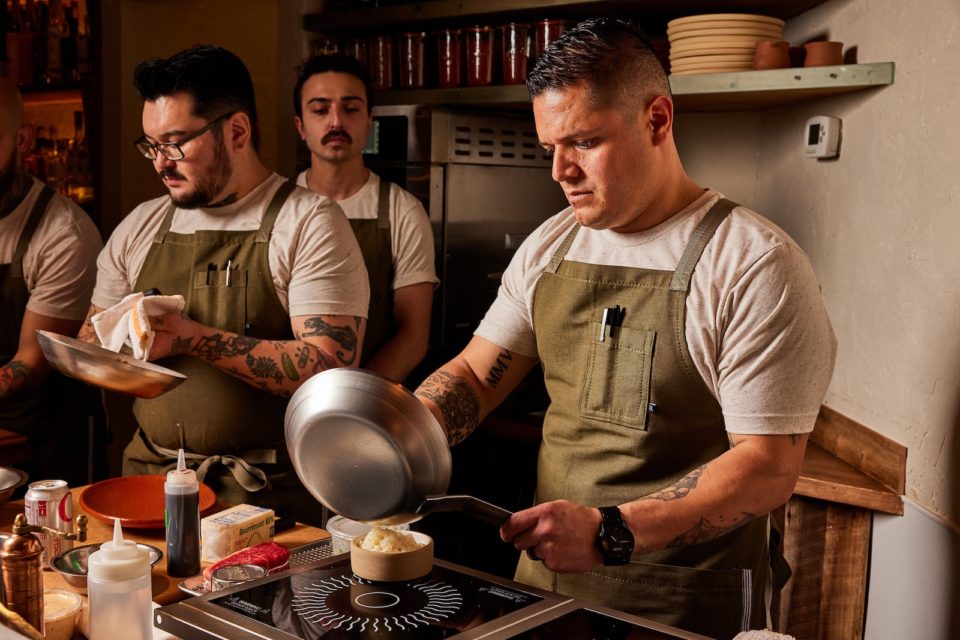 A chef in an olive green apron moves food from a pan into a bowl while other chefs look on.