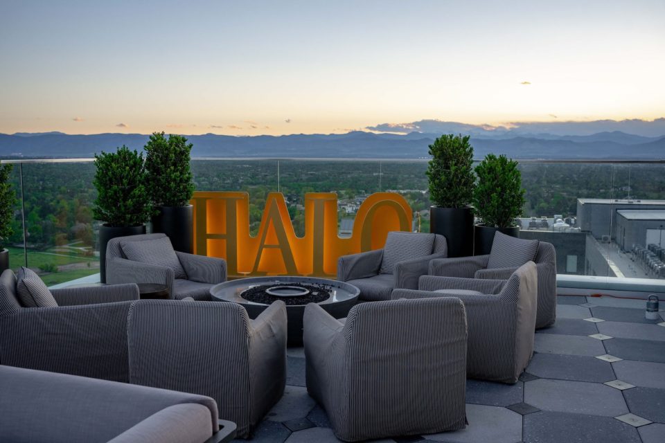 A group of outdoor lounge chairs surround a fire pit with a slgn that reads Halo on a rooftop patio with sunset over mountains in the background.