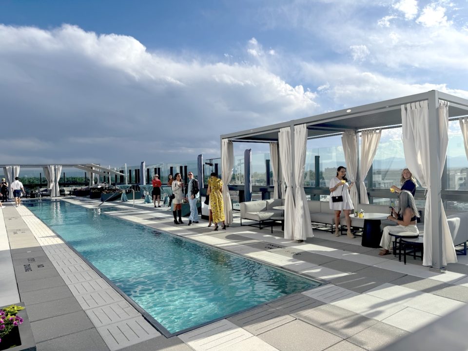 A long, narrow swimming pool surrounded by patio furnishings and bar customers of a rooftop bar with clouds and mountains in the background.