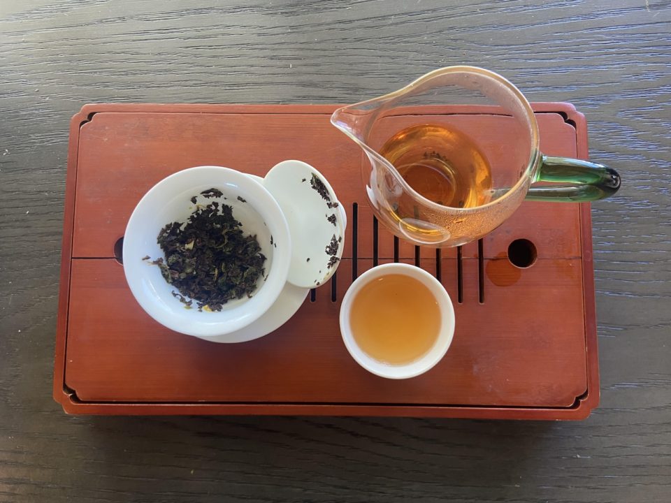 A Japanese tea tray with a glass tea pot, ceramic rea cup, and tea strainer with tea leaves.