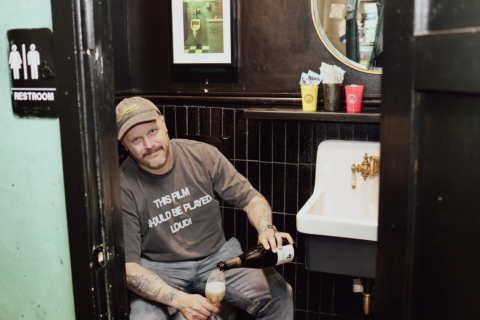Yacht Club co-owner McLain Hedges sitting on the toilet pouring a glass of wine.