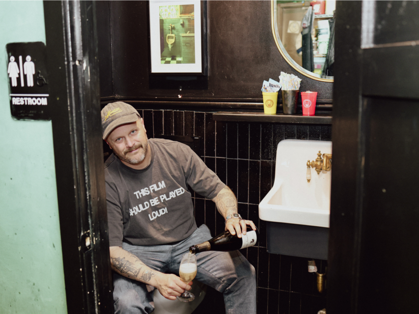 Yacht Club co-owner McLain Hedges sitting on the toilet pouring a glass of wine.