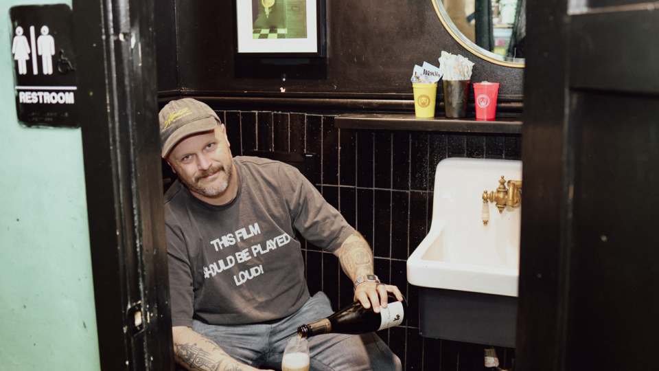 Yacht Club co-owner McLain Hedges sitting on the toilet pouring a glass of wine.