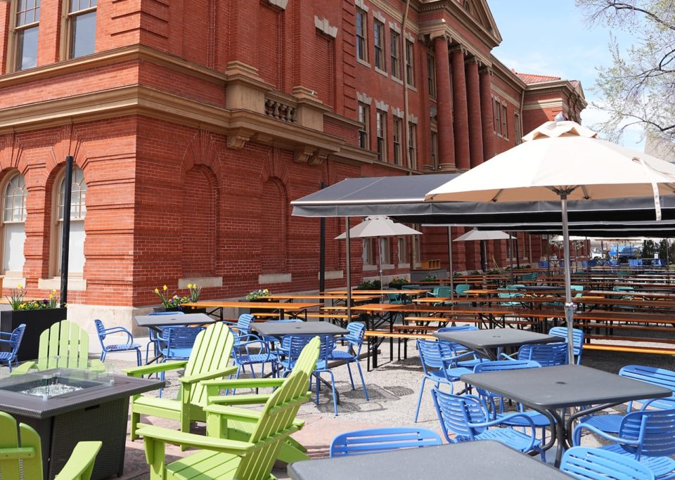 Outdoor chairs, tables, and umbrellas cover the patio of a large red-brick building. 